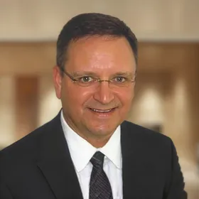 Smiling man with short dark hair and wire-frame glasses, wearing a dark suit and striped tie.