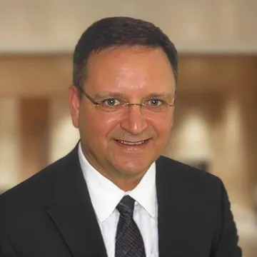 Smiling man with short dark hair and wire-frame glasses, wearing a dark suit and striped tie.