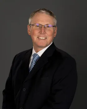 Blair Schadler, smiling, wearing glasses and a dark suit with a blue patterned tie.
