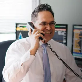 Joseph Maas, smiling, on the phone at his desk in a white dress shirt and tie.